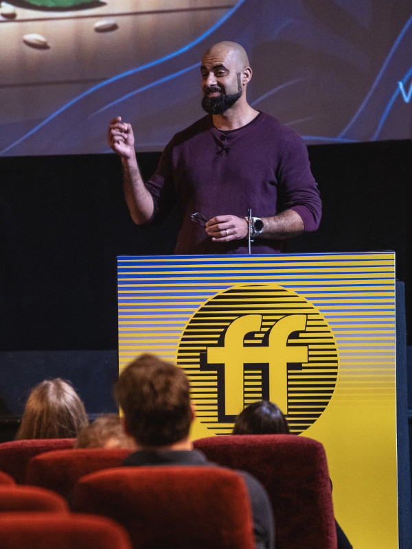 Asim Hussain is behind the podium during his talk, with people watching him from the seats