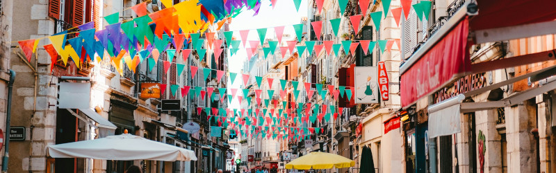 A festive street in a city, with multi-coloured bunting hanging across the street from buildings, in a photo by Tom Sekula from Unsplash